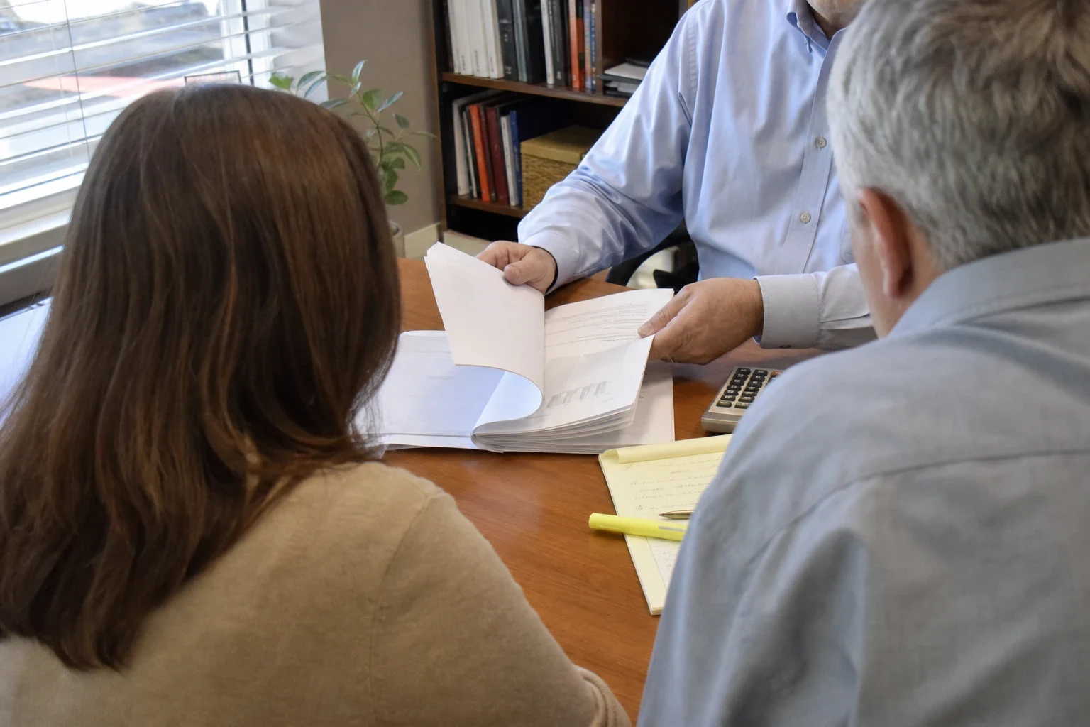 Agent in a blue shirt turns the pages of a bound document for a couple seated across a wooden desk, with a highlighter, notepad and calculator nearby and a bookshelf behind