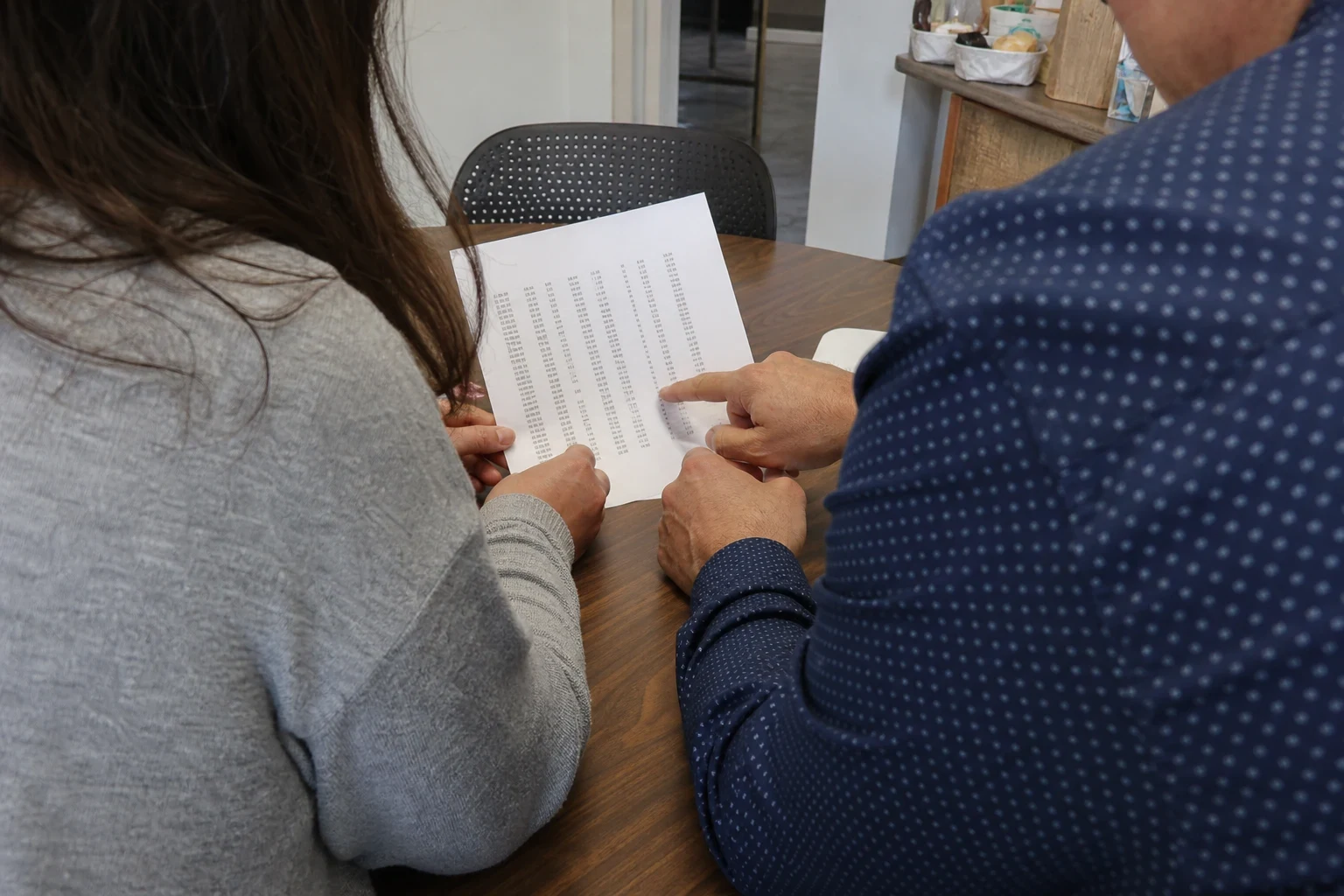 Two people seated at a wooden table reviewing a printed page of figure columns, one pointing at the numbers, in a small storefront office.