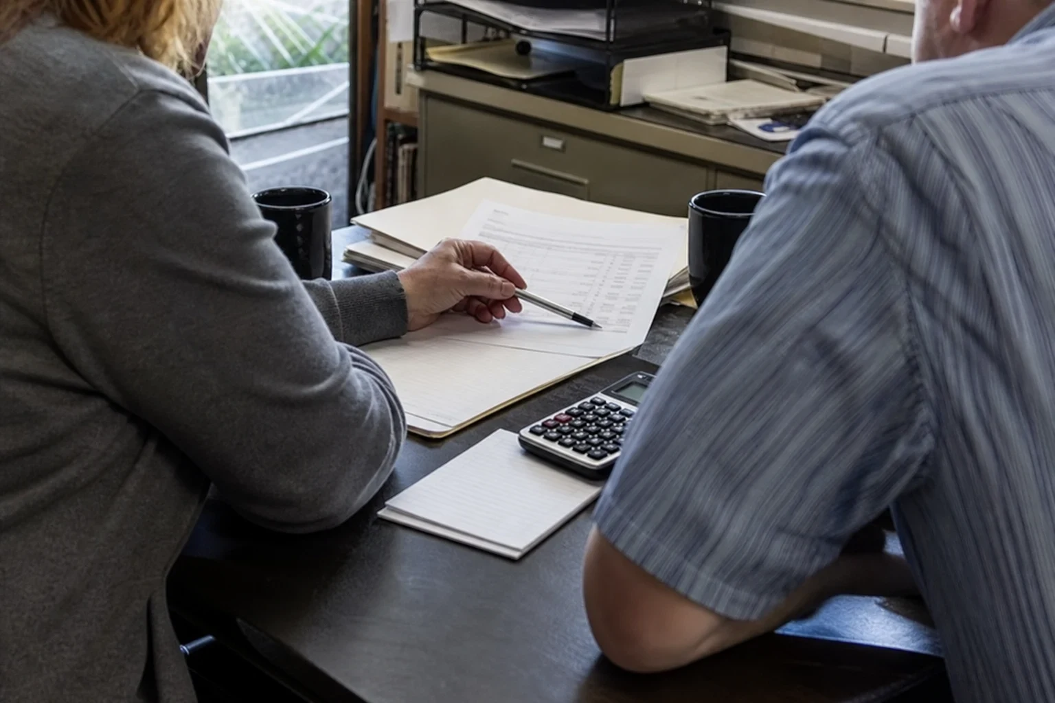 Two people at an office desk reviewing a printed statement with a pen, beside a calculator, notepad, coffee mugs and a filing cabinet.